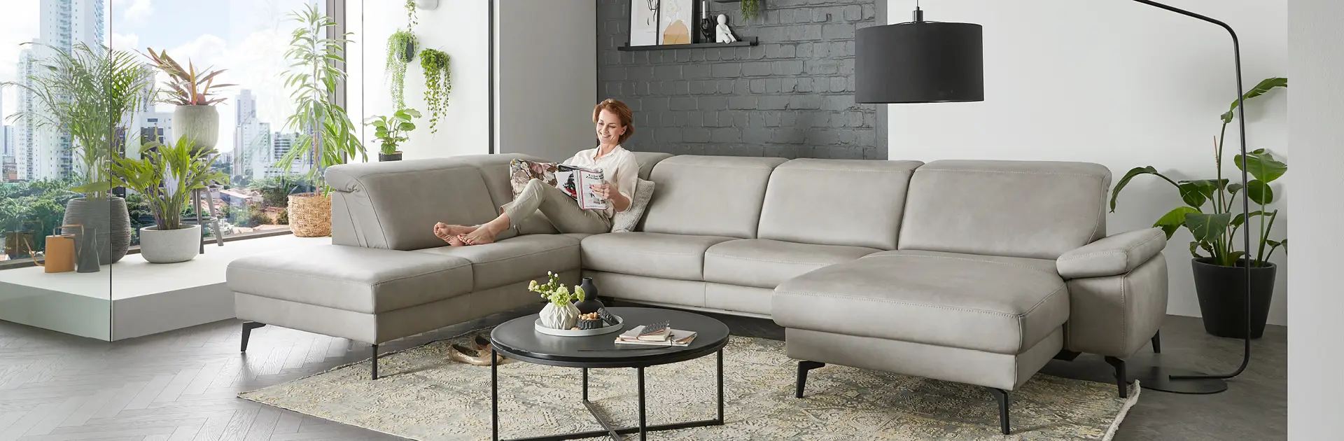 Großes modernes Ecksofa in Hellgrau mit Frau beim Lesen, umgeben von Zimmerpflanzen und Stadtblick durch große Fensterfront